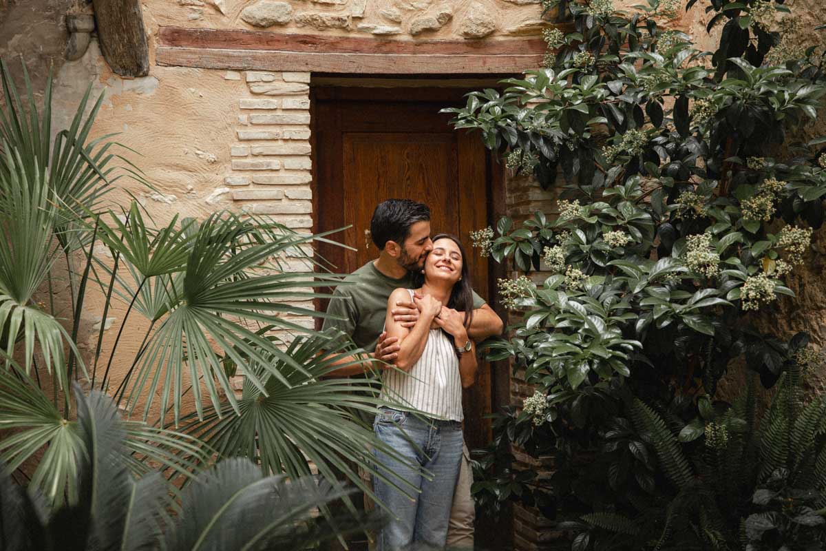 Pareja abrazándose en un rincón con plantas y una puerta antigua de Bocairent durante su preboda